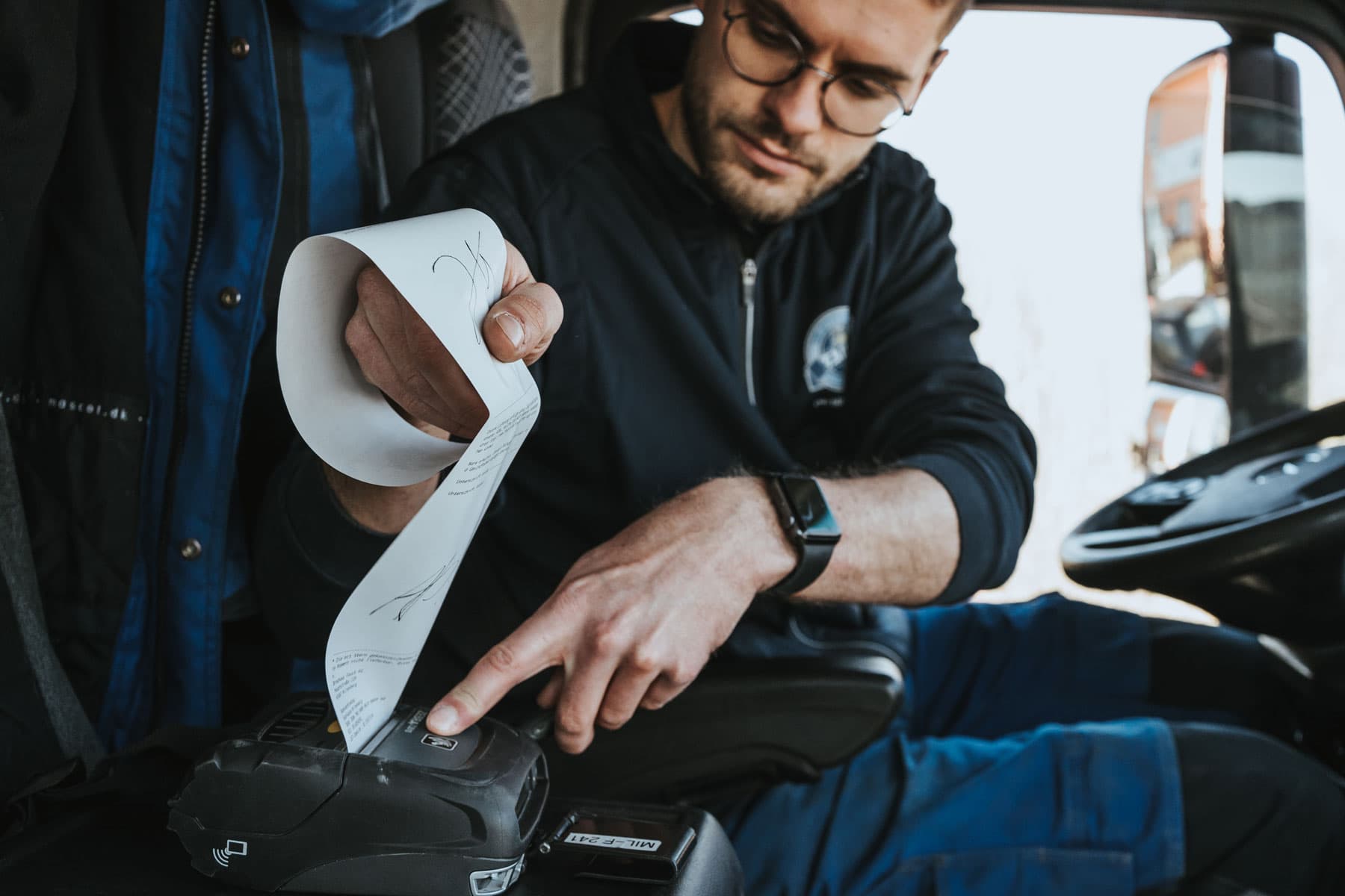 Ein%20Mann%20sitzt%20in%20einem%20Fahrzeug%20und%20bedient%20einen%20mobilen%20Drucker%2C%20w%E4hrend%20er%20einen%20frisch%20gedruckten%20Beleg%20entgegenzieht.%20Eine%20weitere%20Person%20steht%20daneben%20und%20h%E4lt%20das%20Papier%20fest.