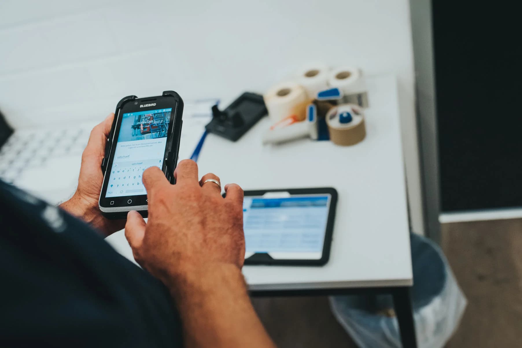 Mitarbeitende%20erfassen%20Daten%20mit%20einem%20robusten%20mobilen%20Ger%E4t%20in%20einer%20Logistik-%20oder%20Lagerumgebung.%20Im%20Hintergrund%20liegen%20ein%20Tablet%20und%20Barcode-Etiketten%20auf%20einem%20Schreibtisch