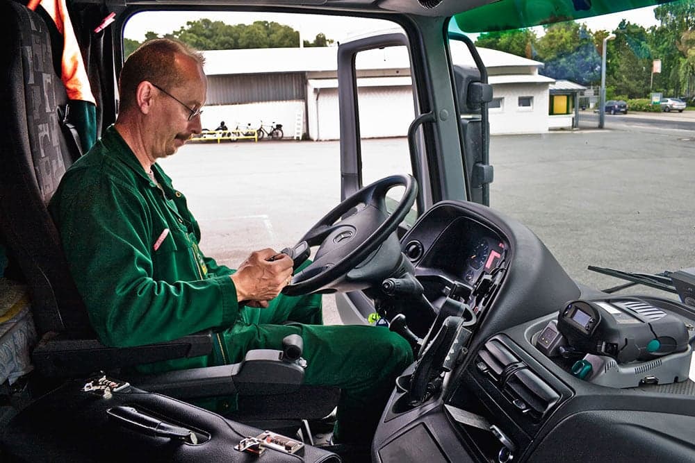 Ein%20Fahrer%20sitzt%20in%20der%20Kabine%20eines%20Lastwagens%20und%20bedient%20ein%20mobiles%20Ger%E4t%2C%20das%20auf%20dem%20Armaturenbrett%20liegt.%20Er%20tr%E4gt%20einen%20gr%FCnen%20Arbeitsanzug%20und%20konzentriert%20sich%20auf%20die%20Eingabe.