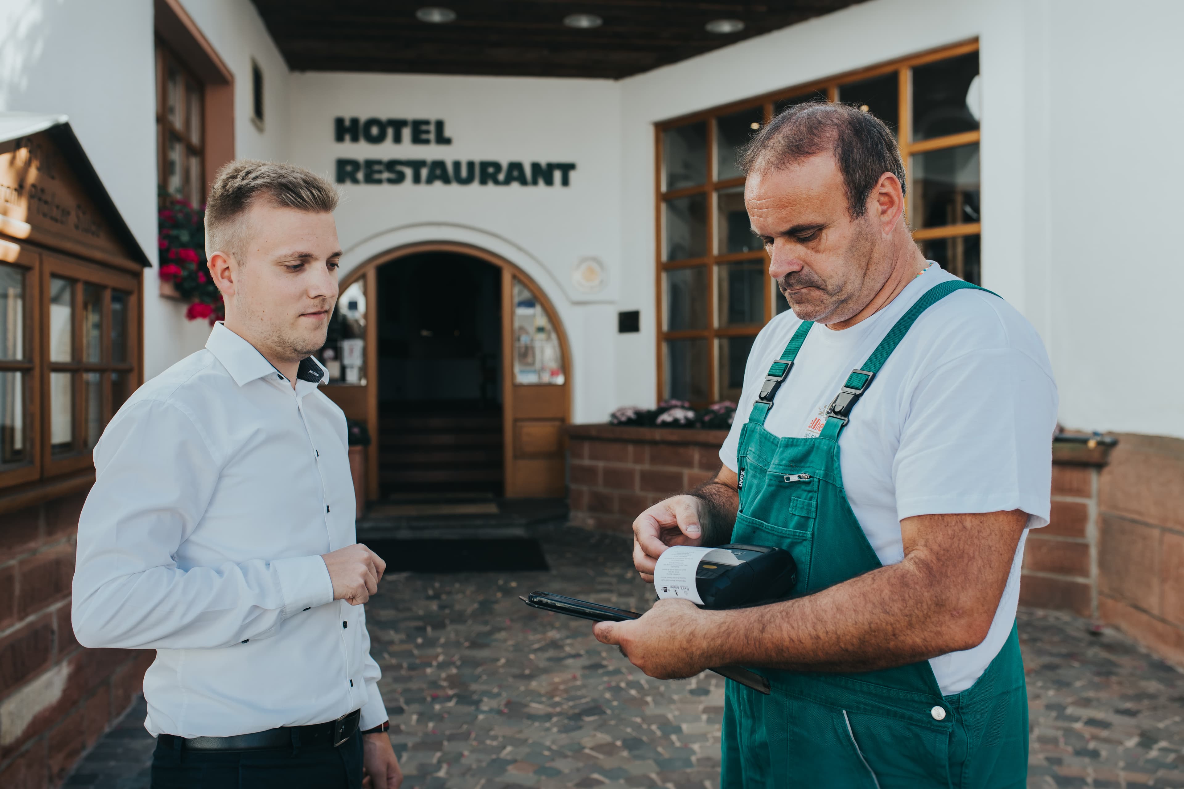 Zustellmitarbeiter%20in%20gr%FCner%20Latzhose%2C%20der%20ein%20mobiles%20Handger%E4t%20zur%20Lieferbest%E4tigung%20vor%20einem%20Hotelrestaurant%20benutzt%2C%20w%E4hrend%20ein%20Mann%20im%20wei%DFen%20Hemd%20zuschaut.