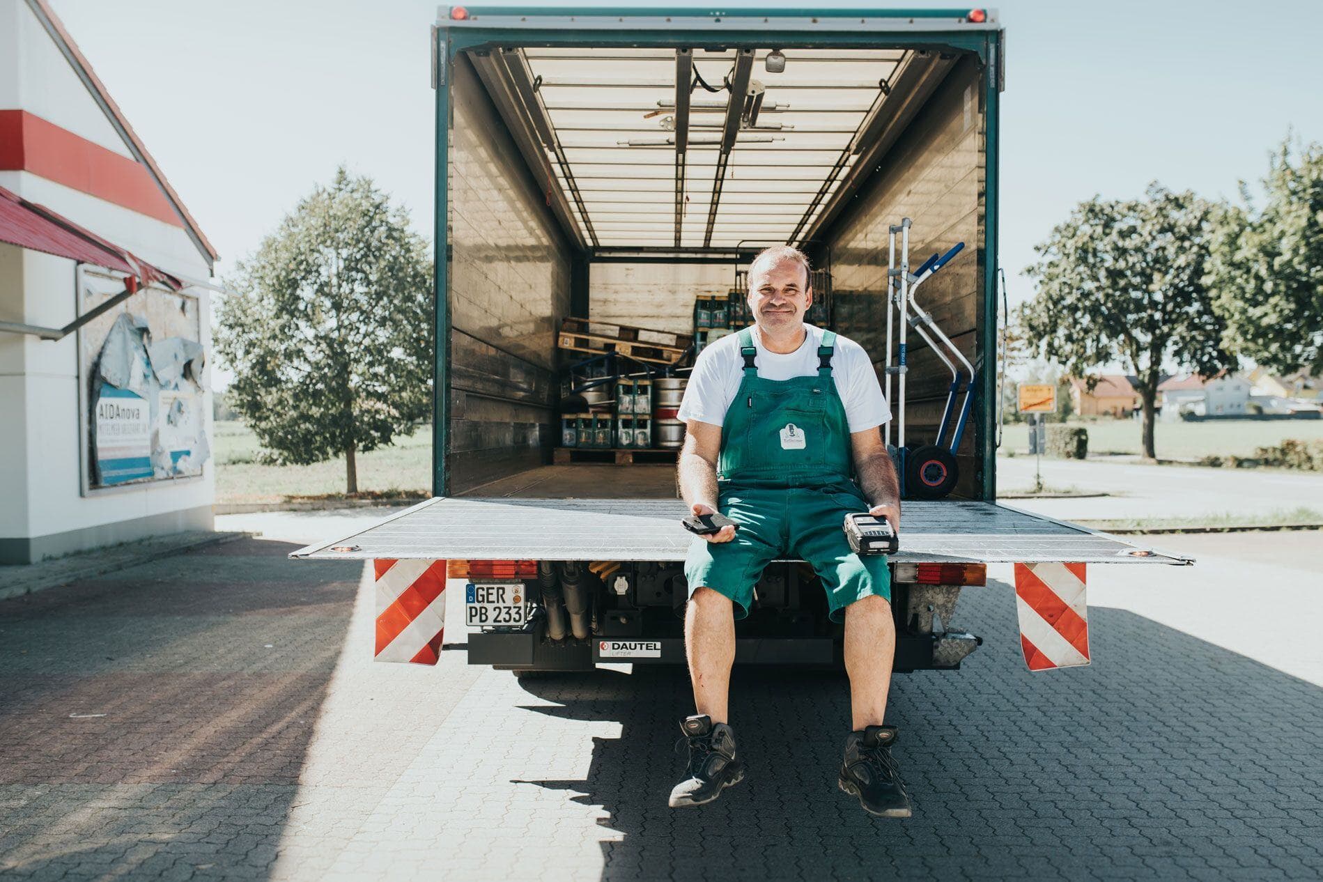 Ein%20Zusteller%20in%20gr%FCnem%20Overall%20sitzt%20l%E4chelnd%20auf%20der%20Ladebordwand%20eines%20LKWs%20und%20h%E4lt%20Handheld-Ger%E4te.