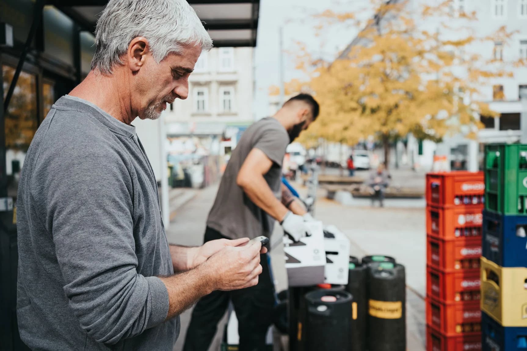 Ein%20Lieferant%20in%20einem%20grauen%20Pullover%20pr%FCft%20ein%20elektronisches%20Handger%E4t%2C%20w%E4hrend%20ein%20Kollege%20im%20Hintergrund%20gestapelte%20Getr%E4nkekisten%20auf%20einer%20Stadtstra%DFe%20entl%E4dt.